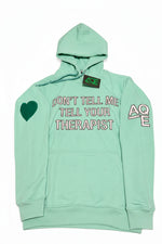 A mint green pullover hoodie with a front kangaroo pocket, featuring white text that reads 'DON'T TELL ME TELL YOUR THERAPIST' across the chest, a green heart on the right sleeve in support of mental health awareness and white 'AQE' text on the left sleeve 