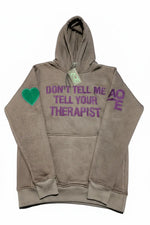 A taupe-grey pullover mental health awareness hoodie with purple embroidered text reading 'DON'T TELL ME TELL YOUR THERAPIST' across the chest, a green heart patch on the right sleeve, and purple 'AQE' embroidery on the left sleeve photographed on a plain white background.